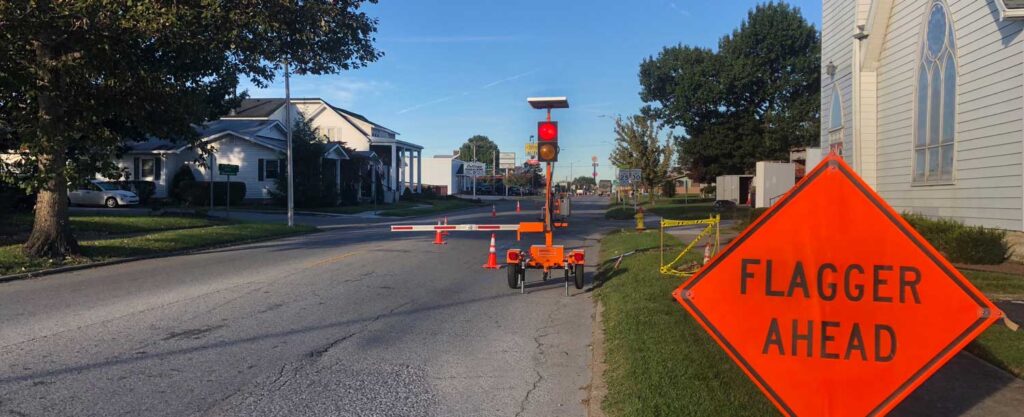 Automated flagger vs human flagger, device holding one end of a street lane closure with advance flagger ahead signing